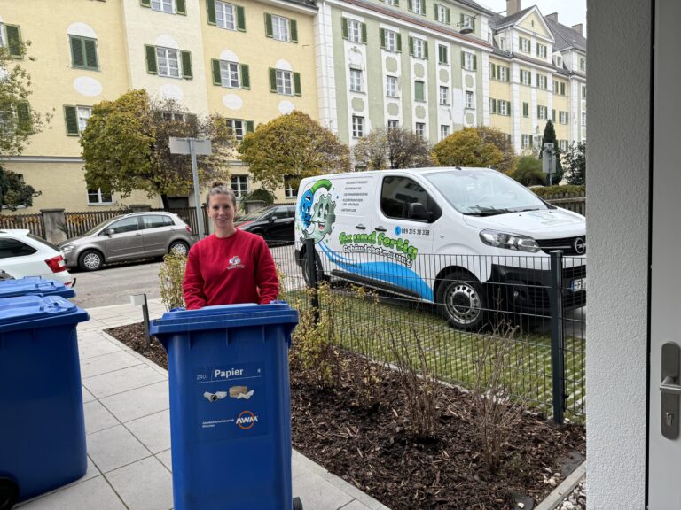 Ein Hausmeister stellt die blauen Papiertonnen für die Müllabfuhr bereit. Im Hintergrund steht das Auto des Hausmeisterservices, ein Lieferwagen mit dem Logo von fix und fertig Gebäudebetreuung. Im Hintergrund sind die Häuser einer Wohnanlage zu sehen.